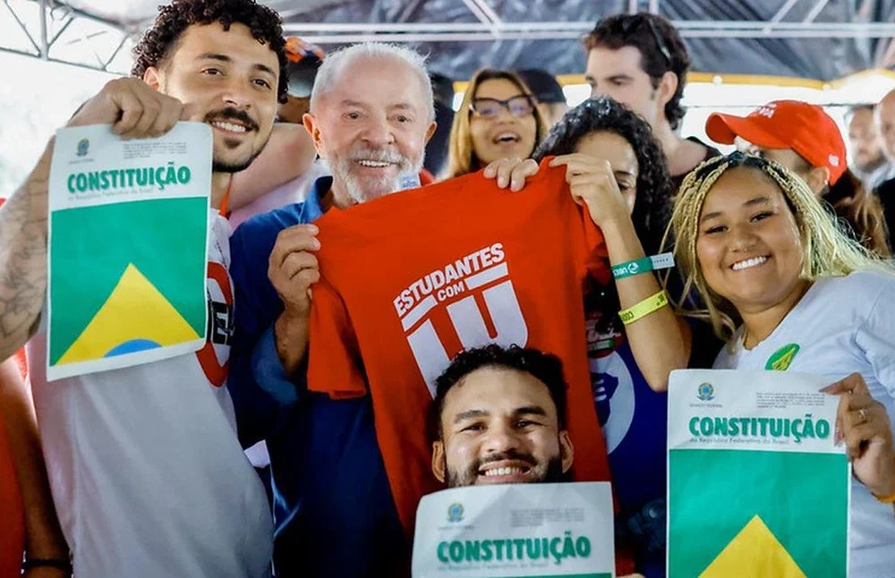Lula em vídeo selfie rodeado por jovens ao fundo, anunciando o programa universitário Pé de Meia